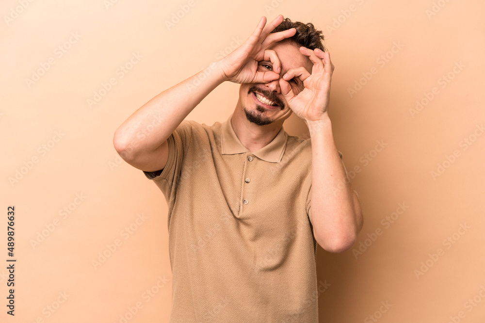 Young caucasian man isolated on beige background showing okay sign over eyes Young caucasian man isolated on beige background showing okay sign over eyes
