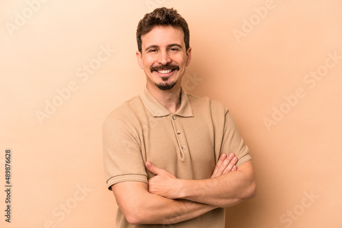 Young caucasian man isolated on beige background who feels confident, crossing arms with determination.