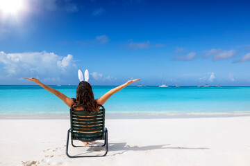 Easter travel concept with a happy woman with bunny ears sitting on a tropical beach with outstretched arms