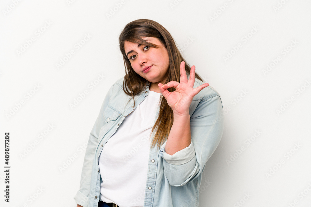 Young caucasian overweight woman isolated on white background winks an eye and holds an okay gesture with hand. Young caucasian overweight woman isolated on white background winks an eye and holds an okay gesture with hand.