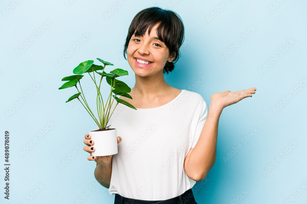 Young hispanic woman holding a plant isolated on blue background showing a copy space on a palm and holding another hand on waist. Young hispanic woman holding a plant isolated on blue background showing a copy space on a palm and holding another hand on waist.