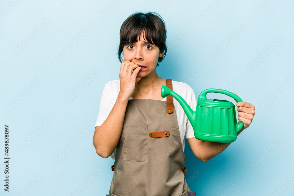 Young hispanic gardener woman holding a watering can isolated on blue background biting fingernails, nervous and very anxious. Young hispanic gardener woman holding a watering can isolated on blue background biting fingernails, nervous and very anxious.