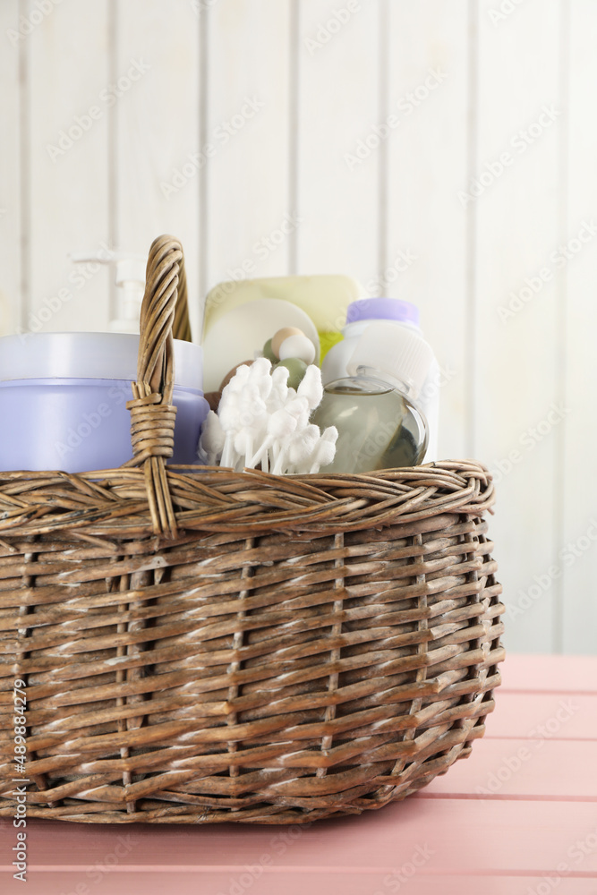 Wicker basket full of different baby cosmetic products and bathing accessories on pink wooden table, closeup