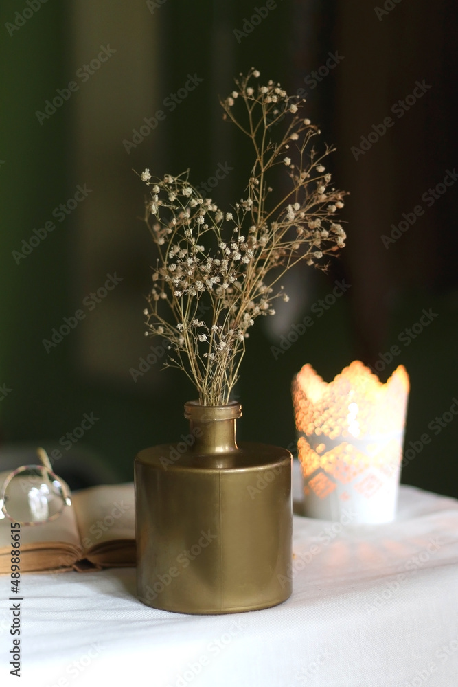 Open book, reading glasses, candle holder with lit candle and vase with gypsophila flowers. Dark background, selective focus.
