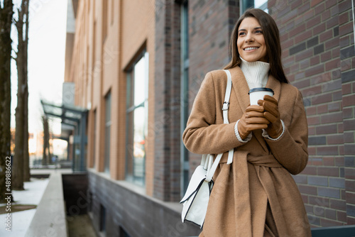 Joyful woman in a brown coat with a paper cup of coffee on the street