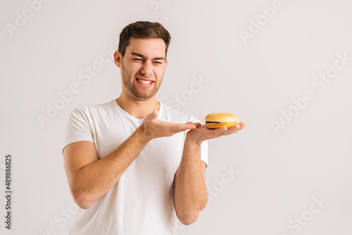 Portrait of dissatisfied young man with disgust pointing to bad burger on white isolated background. Studio shot of handsome bearded male holding in hands unhealthy delicious hamburger.