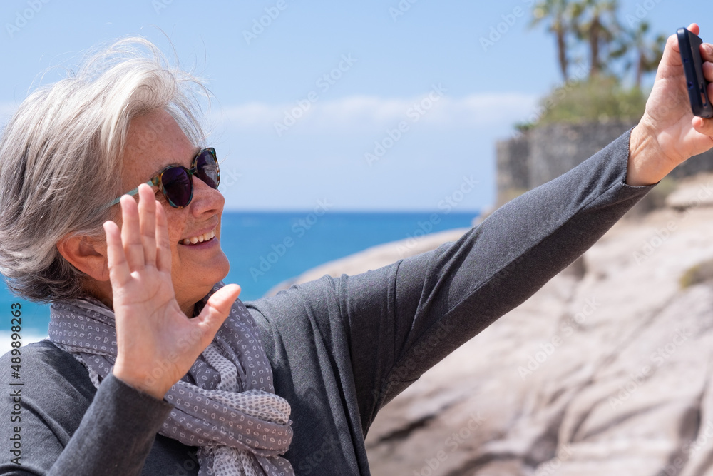 Smiling senior woman wave to camera having video call on mobile phone outdoor at sea vacation. Happy elderly female standing on the beach talk using modern technologies and wireless connection.