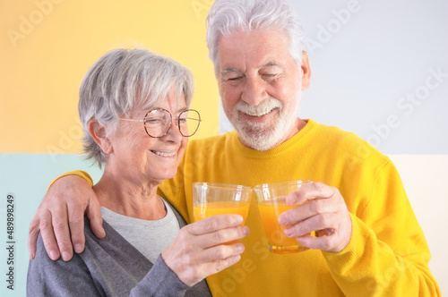 Happy caucasian senior couple standing over isolated colorful background hugging holding a glass of healthy orange juice. Elderly people smiling carefree enjoying retirement, healthy lifestyle