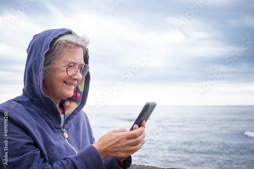 Attractive mature senior pensioner woman enjoying retirement at sea. Caucasian elderly female standing on the beach in a cloudy day using mobile phone enjoying tech and social. Horizon over water
