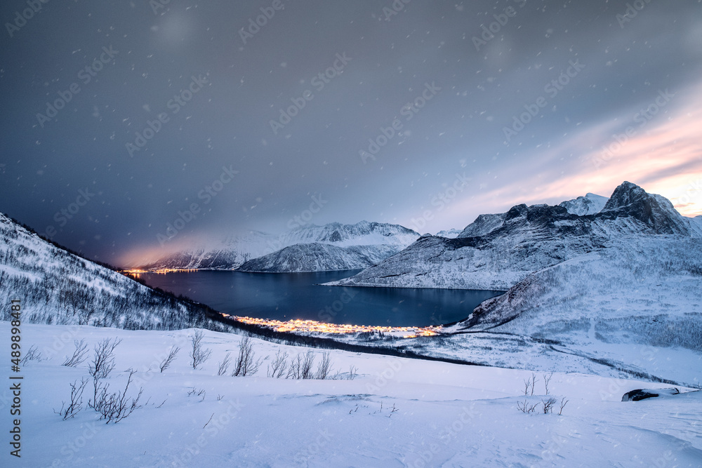 Snow covered mountain range with illuminated town on coastline in blizzard Snow covered mountain range with illuminated town on coastline in blizzard