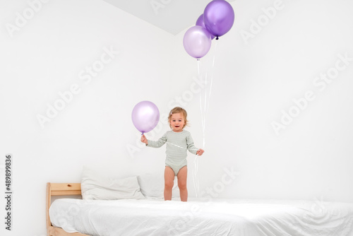 Cute funny caucasian blonde baby girl,toddler of 1,2 year old playing on bed with big purlple balloons of very peri trendy color on white background in modern scandinavian interior at home.Copy space photo
