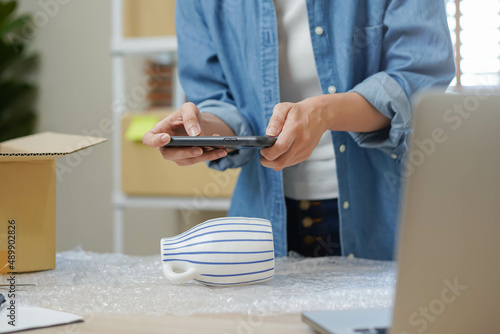 Small business entrepreneur SME, asian young woman owner packing product box, delivery, using smartphone take a photo, checking parcel before send order to customer. Merchant, work online, ecommerce.