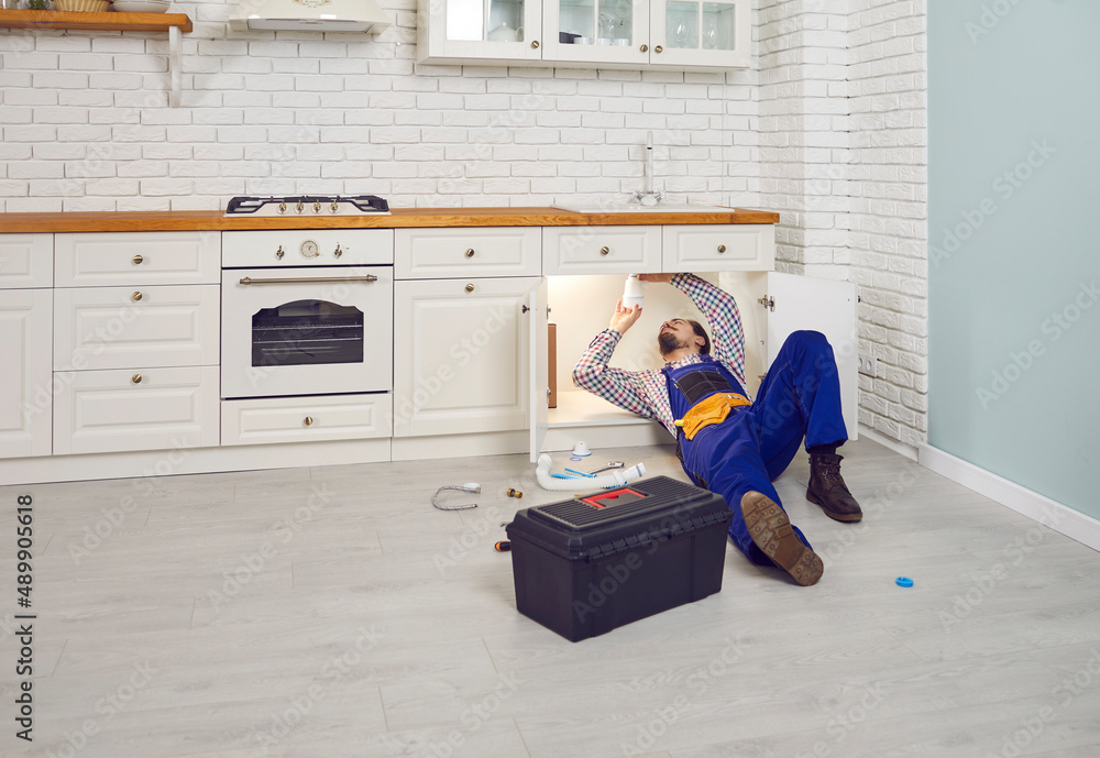 Plumber cleaning out the clogged drain. Young handyman in an overall uniform lying down on the floor under the sink in a modern kitchen interior and fixing some problems with the pipes