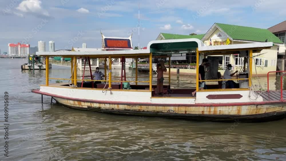 BANGKOK, THAILAND - Circa November, 2021: Boat on Chao Phraya River with passengers enters the pier in slow motion during a sunny day with blue sky and a riverside restaurant in the background. BANGKOK, THAILAND - Circa November, 2021: Boat on Chao Phraya River with passengers enters the pier in slow motion during a sunny day with blue sky and a riverside restaurant in the background.