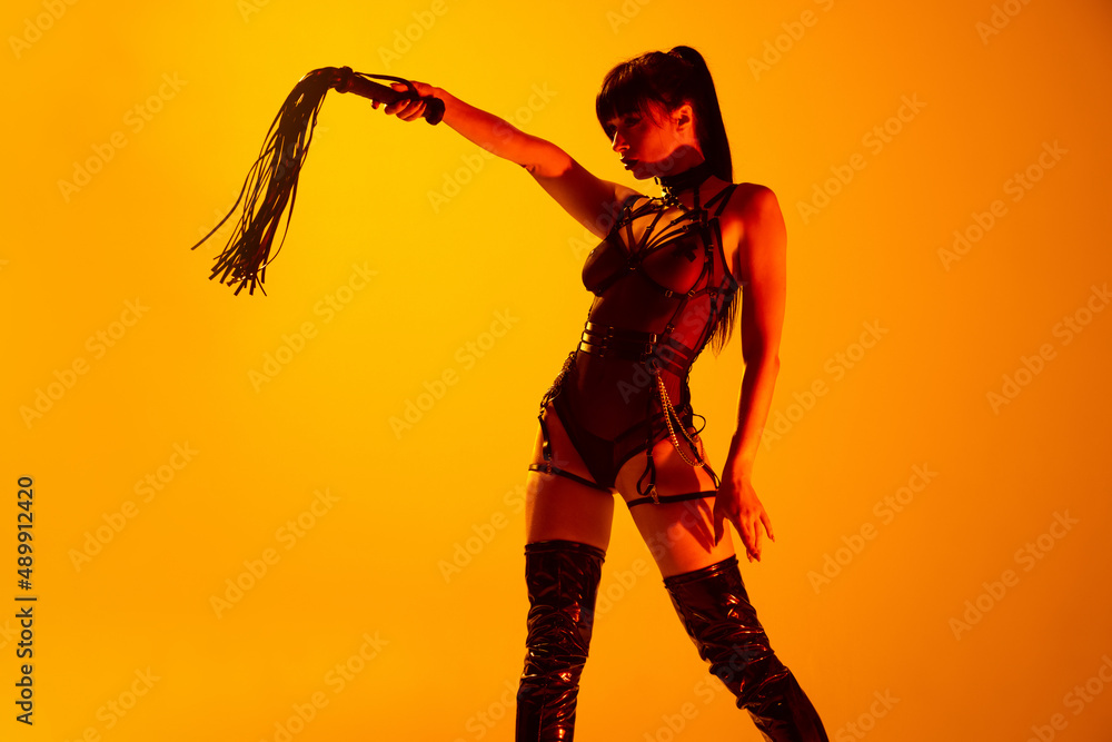 Obraz premium Studio shot of sexy young woman wearing leather lingerie posing isolated on yellow background in neon light. Concept of beauty, love