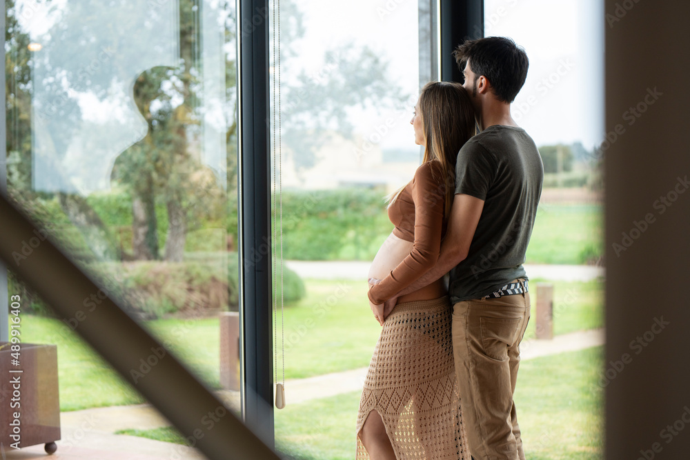 Young man hugging her pregnant woman and looking out the window 