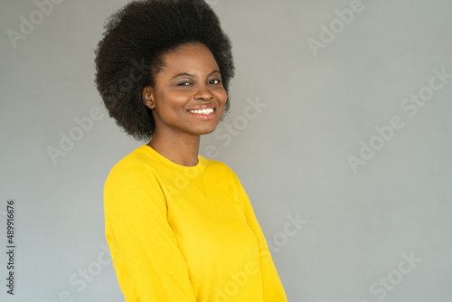 Positive young black woman with cute cheery smile looking at camera, wear casual yellow sweater. Happy african american girl with curly hairstyle cheerful and satisfied isolated over gray studio wall