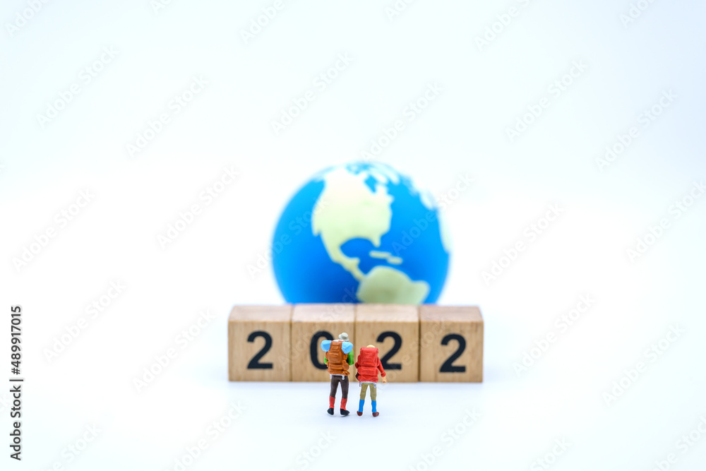 2022 New Year and Travel Concept. Closeup of two of traveler miniature figures with backpack standing with stack of wooden number block and mini world ball on white background.