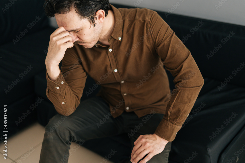High-angle view of worried young man sitting alone feeling sad worry regret or fear, depressed and desperate in sofa at home. Suffering emotional pain and unhappy, mental health. High-angle view of worried young man sitting alone feeling sad worry regret or fear, depressed and desperate in sofa at home. Suffering emotional pain and unhappy, mental health.