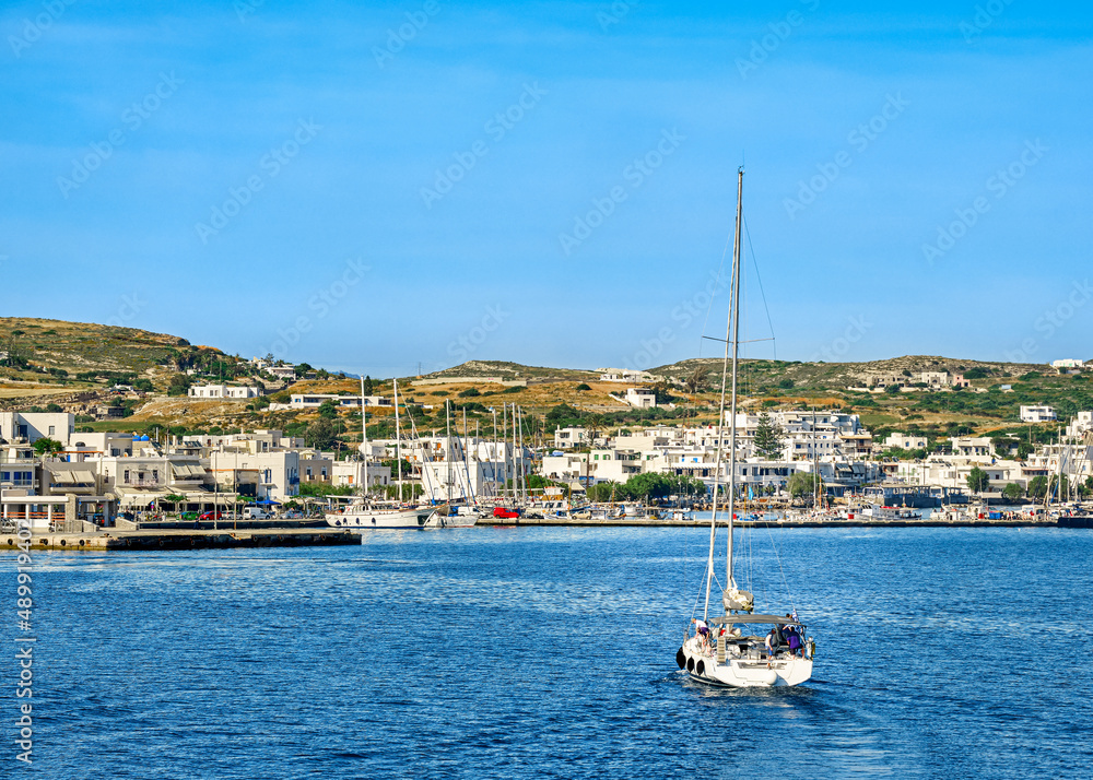 Sailboat going to port of Greek island. Yachts and fishing boats in harbor at sunny summer day. Whitewashed houses on green hills.