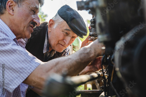 Two caucasian senior men father and son pensioner at the farm repairing the tractor machine in sunny day family bonding generation cooperation and togetherness concept real people
