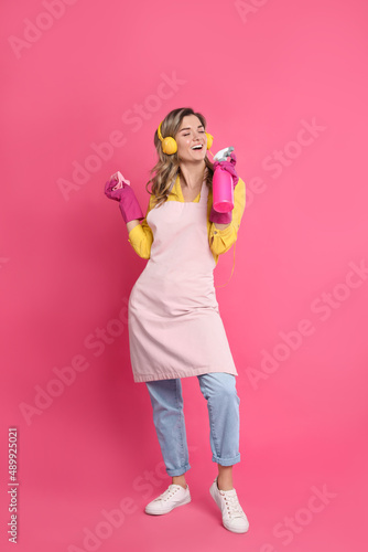 Beautiful young woman with headphones, rag and bottle of detergent singing on pink background © New Africa