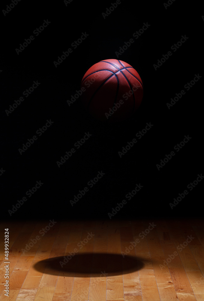 Basketball spot lit suspended in air against a black background above a hardwood maple court floor