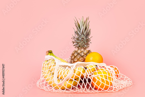 bananas, oranges and pineapple in a shopping bag, string bag lie on an isolated pastel pink background with copyspace. Food conept. Nutrition concept. Eco activism concept. ecology concept.