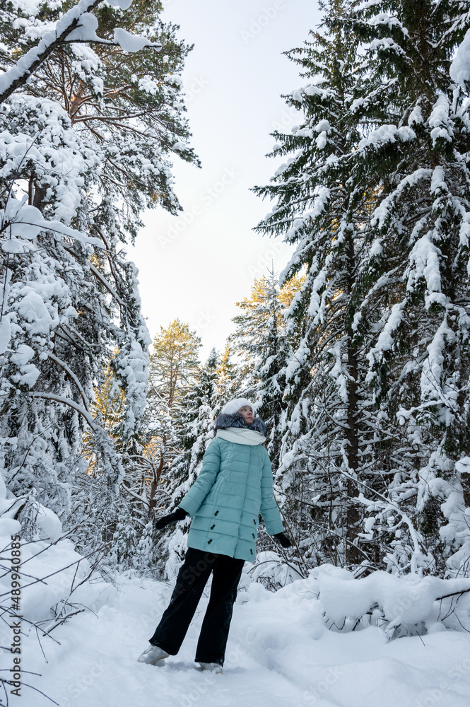 pretty young girl walks in winter forest among tall pines covered with snow. Girl is wearing warm jacket, white hat and . scarf. Clothes for walking in winter