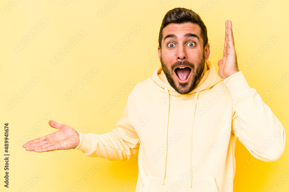 Young caucasian man isolated on yellow background holds copy space on a palm, keep hand over cheek. Amazed and delighted. Young caucasian man isolated on yellow background holds copy space on a palm, keep hand over cheek. Amazed and delighted.