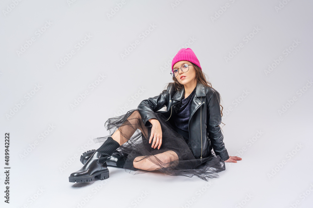 stylish teenage girl with dark hair, dressed in fashionable clothes, a leather jacket and a pink hat and glasses on white wall. full growth. concept of youth and clothing. stylish teenage girl with dark hair, dressed in fashionable clothes, a leather jacket and a pink hat and glasses on white wall. full growth. concept of youth and clothing.