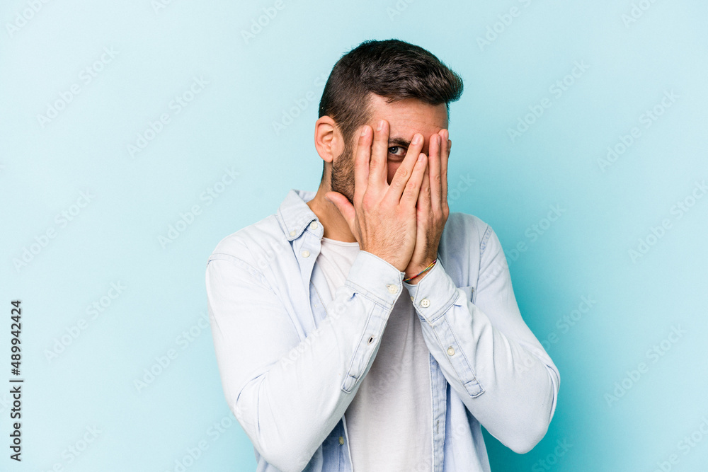 Young caucasian man isolated on blue background blink at the camera through fingers, embarrassed covering face. Young caucasian man isolated on blue background blink at the camera through fingers, embarrassed covering face.
