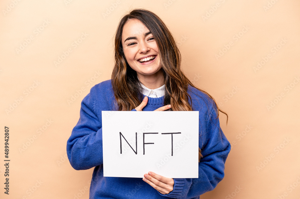 Young caucasian woman holding a NFT placard isolated on beige background laughs out loudly keeping hand on chest. Young caucasian woman holding a NFT placard isolated on beige background laughs out loudly keeping hand on chest.