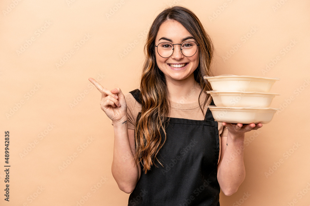 Young caucasian woman holding tupperware isolated on beige background smiling and pointing aside, showing something at blank space. Young caucasian woman holding tupperware isolated on beige background smiling and pointing aside, showing something at blank space.
