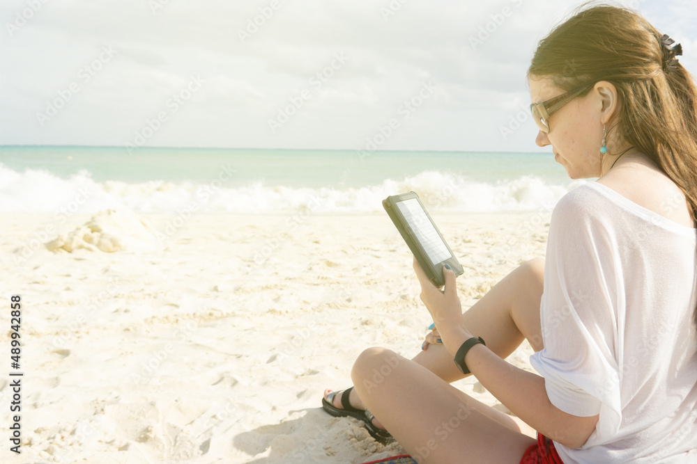 A girl on a deserted sandy beach reads an e-book. The girl reads on the background of the sea.