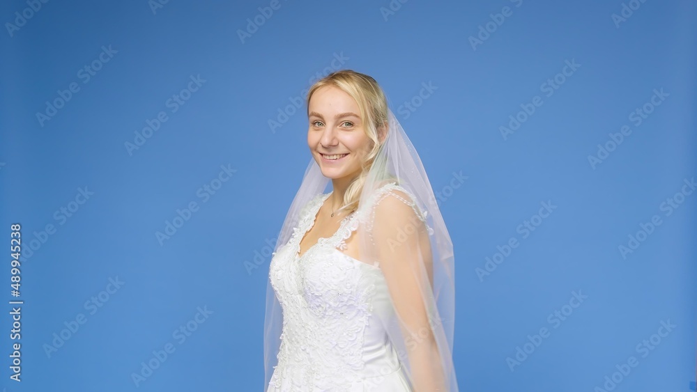 Young beautiful blonde smiling in a wedding white dress and veil on a blue background. The girl looks into the camera. Wedding Young beautiful blonde smiling in a wedding white dress and veil on a blue background. The girl looks into the camera. Wedding