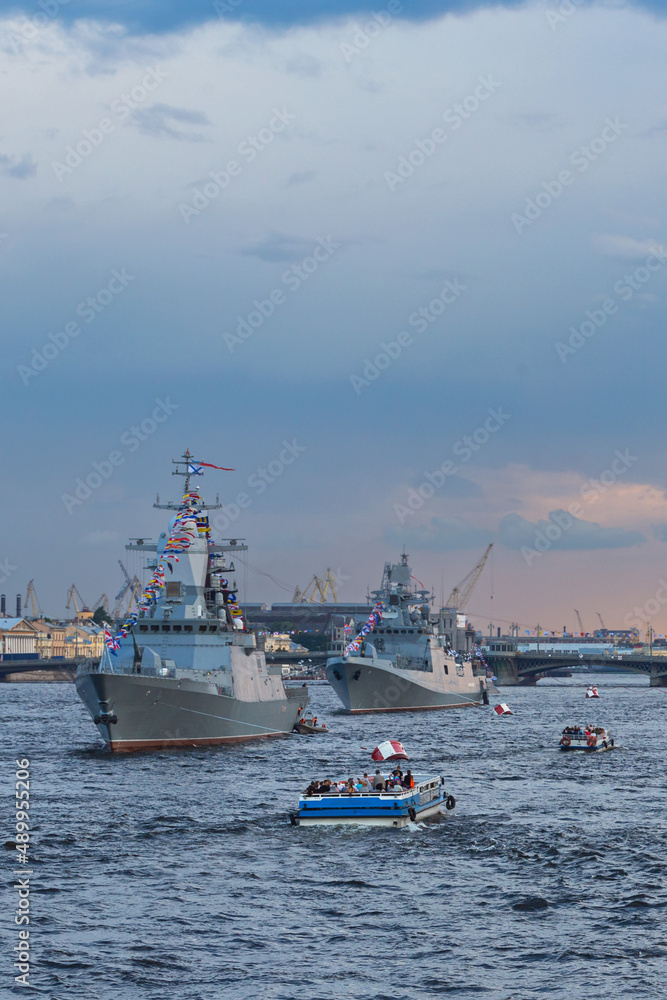 small boats with tourists inspect warships on the Neva in the city center against the blue evening sky