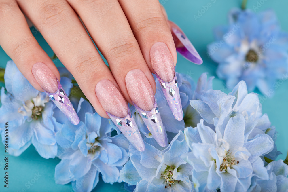 Fototapeta premium Hand with long artificial french manicured nails decorated with gemstones and blue flowers. Fashion and stylish manicure.