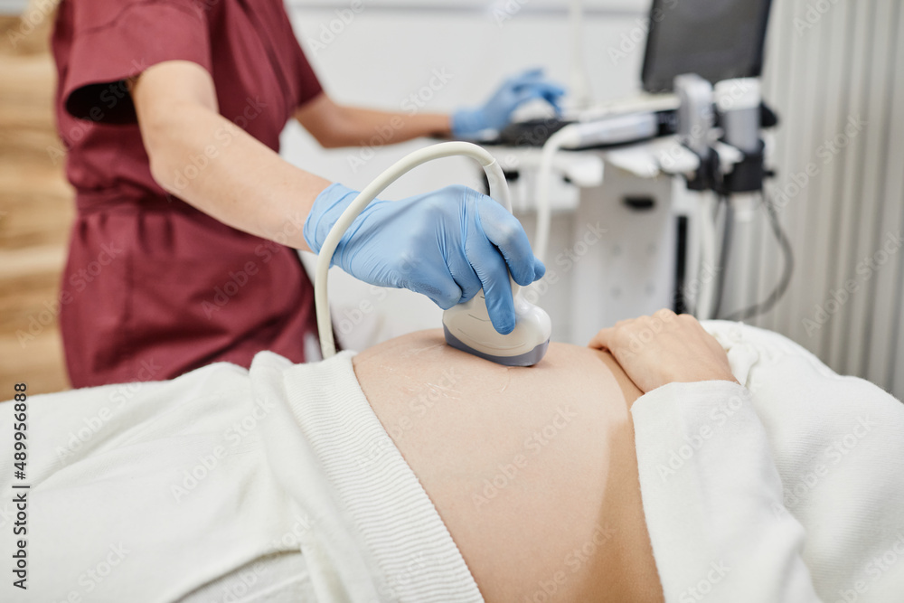 Close up of female doctor holding ultrasound transducer to pregnant belly while examining expectant young woman in clinic, copy space Close up of female doctor holding ultrasound transducer to pregnant belly while examining expectant young woman in clinic, copy space