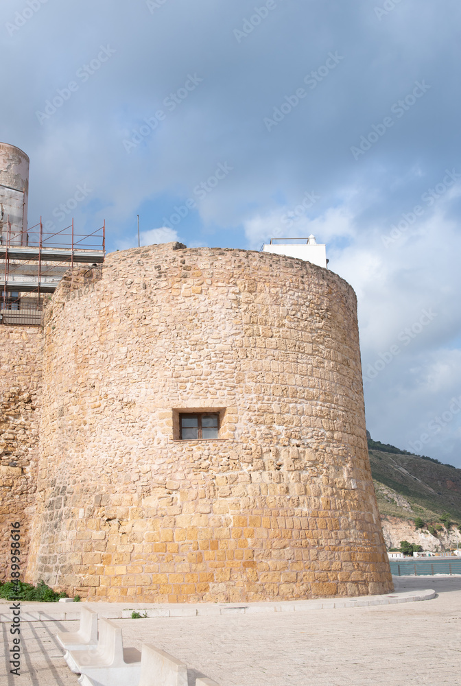 In Castellamare di golfo, in Sicily, stands an old castle from the 14th century on the coast. Part of the fort can be seen in portrait format against a blue sky.