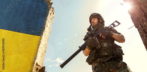Ukrainian Soldier military in the war with a weapon in his hands. The flag of Ukraine is painted on a brick wall. Relations between Ukraine and Russia