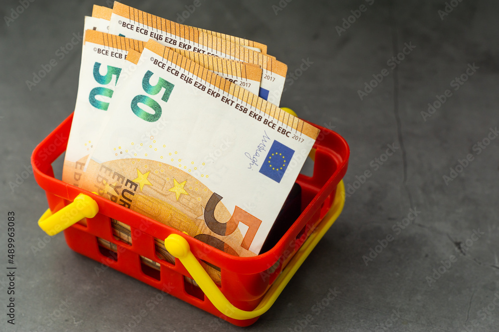 Red shopping basket filled with European Euro banknotes on gray stone background. Raising prices, inflation and expensive life costs concept.