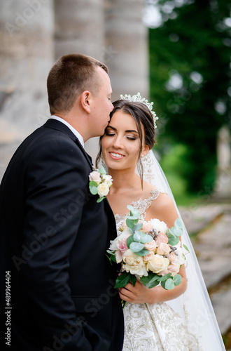 Close view of handsome bridegroom in black suit, standing and kissing wife face while she feeling happy and hugging his during wedding walk outdoor