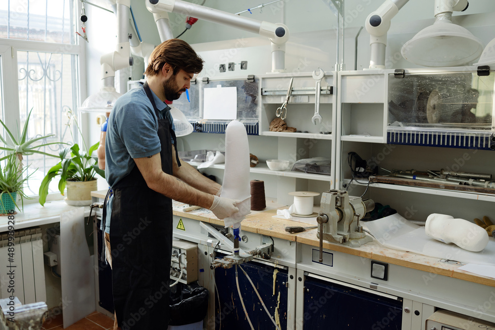 Fototapeta premium Worker of manufactory putting white sheet on workpiece of prosthetic socket to form and fix its shape by workplace