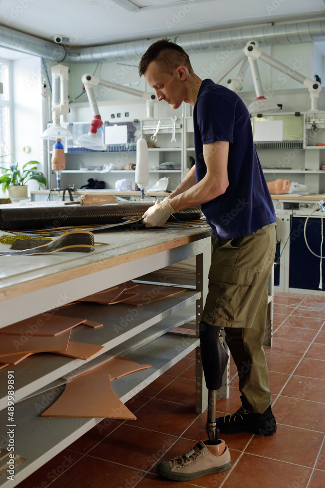 Young man with disability standing by workplace while cutting rolled material necessary in production of prostheses Young man with disability standing by workplace while cutting rolled material necessary in production of prostheses