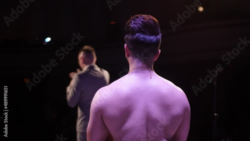 An Indian man without outerwear stands on stage. The bare back of an Indian dancer. Big stage. The host speaks on the microphone. photo