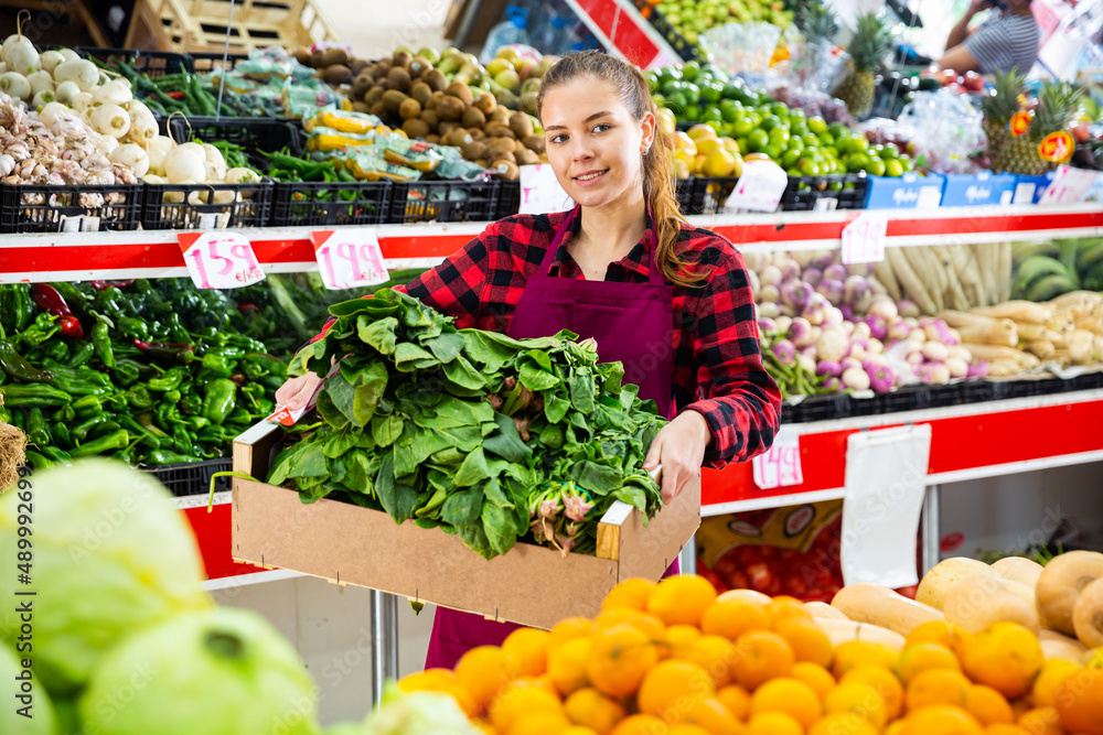 Portrait of a positive young saleswoman near the counter in a store, holding a box of spinach