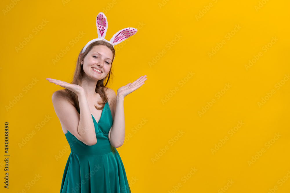 Cheerful young woman with fluffy pink bunny ears, disappointment aside, appreciates holiday tradition, pollution in green dress, stands over yellow studio wall. Seasonal holiday concept. Cheerful young woman with fluffy pink bunny ears, disappointment aside, appreciates holiday tradition, pollution in green dress, stands over yellow studio wall. Seasonal holiday concept.