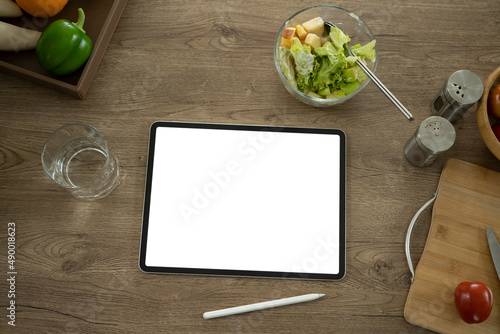 Digital tablet and bowl of healthy vegan salad on wooden table in home kitchen.