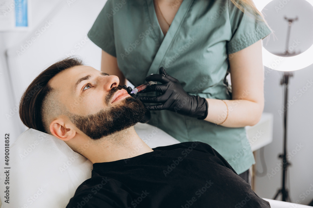 Fototapeta premium Stylish bearded man visiting aesthetic clinic, getting lips filler, closeup. Attractive Man having beauty injection at male spa salon. Anti-aging treatment for men concept.